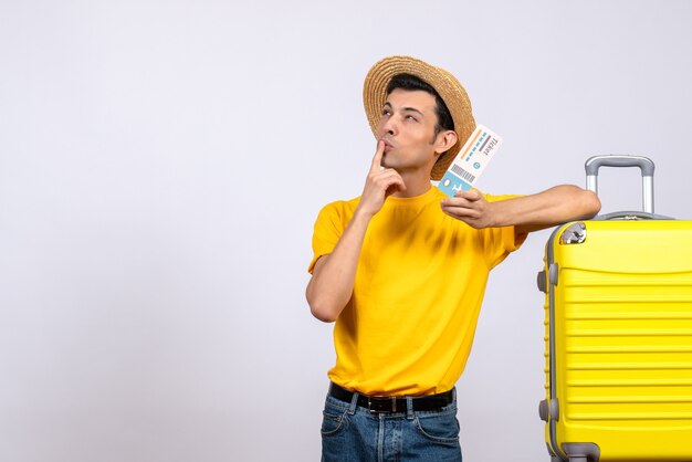 Vue de face jeune touriste debout près de valise jaune tenant un billet pensant à quelque chose