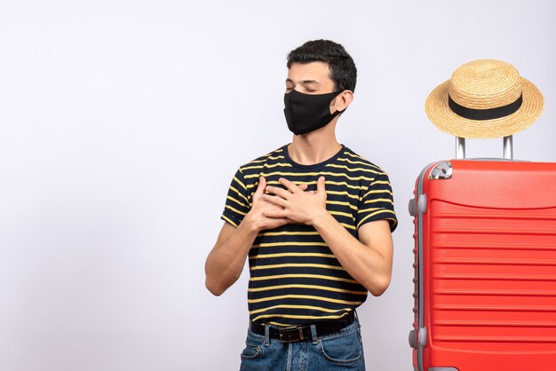 Vue de face jeune touriste gratifié avec masque noir debout près de valise rouge