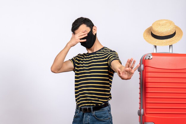 Vue de face jeune touriste avec masque noir debout près de valise rouge fermant les yeux avec la main