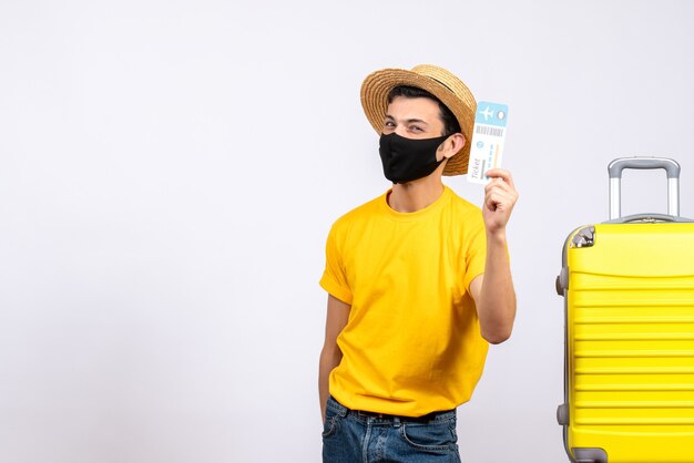 Vue de face jeune touriste en t-shirt jaune debout près de valise jaune tenant un billet de voyage