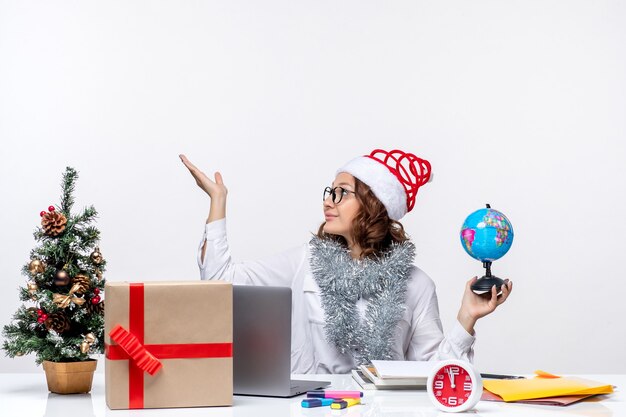 Vue de face jeune travailleur féminin assis devant sa place tenant le globe terrestre sur le sol blanc bureau de travail de bureau de travail de Noël