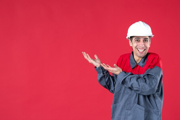 Vue de face d'un jeune travailleur souriant en uniforme avec un casque et montrant quelque chose sur le côté droit sur un mur rouge isolé