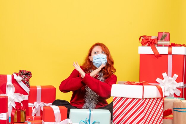 Vue de face jolie femme assise autour de cadeaux en masque sur bureau jaune couleur nouvelle année virus covid- xmas