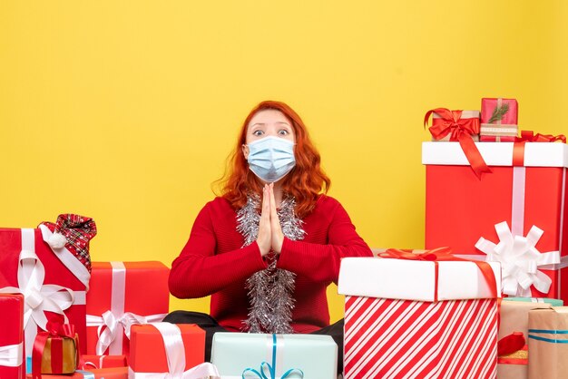 Vue de face jolie femme assise autour de cadeaux en masque priant sur jaune