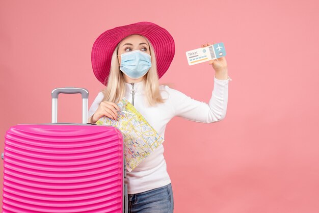 Vue de face jolie femme blonde en masque debout près de valise tenant un billet d'avion