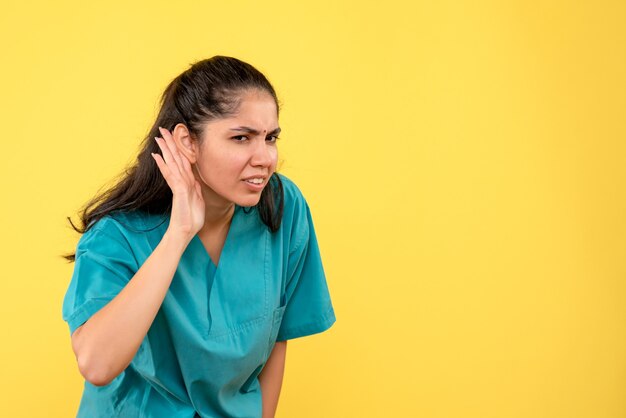 Vue de face de la jolie femme médecin écoutant quelque chose sur le mur jaune