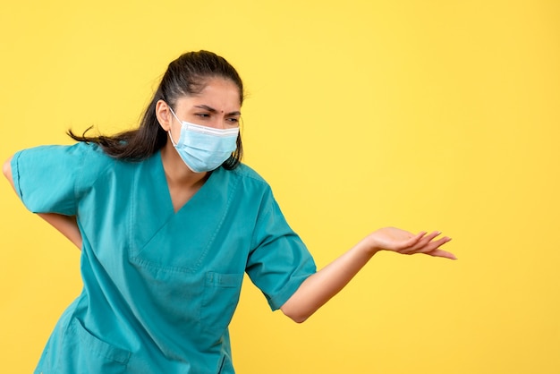 Vue de face de la jolie femme médecin avec masque médical tenant son dos sur le mur jaune