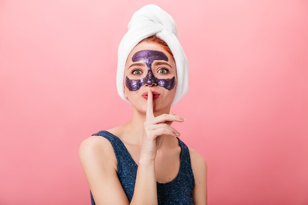 Vue de face d'une jolie femme montrant un signe secret tout en faisant un traitement spa. Photo de Studio de jolie fille avec masque facial isolé sur fond rose.