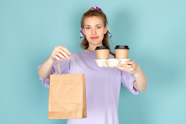 Une vue de face jolie femme en robe-chemise bleue tenant un paquet de papier et des tasses à café sur bleu