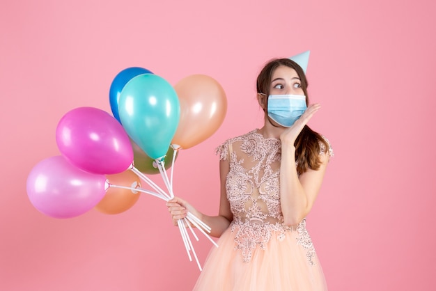 Vue de face jolie fille de fête avec chapeau de fête tenant des ballons colorés regardant à droite