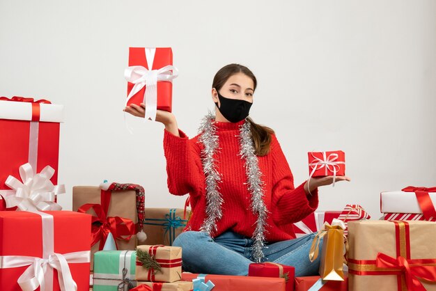 Vue de face jolie fille avec masque noir tenant des cadeaux dans les deux mains assis autour de cadeaux