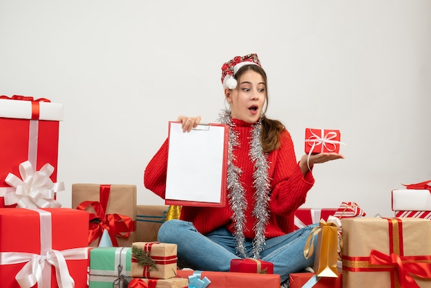 Vue de face jolie fille de Noël avec bonnet de Noel tenant un cadeau et un document assis autour de cadeaux
