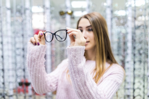 Vue de face de jolie fille en pull blanc essayer des lunettes en magasin sur