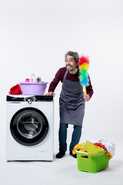 Vue de face joyeuse femme de ménage tenant un plumeau debout près du panier à linge de la machine à laver sur fond blanc