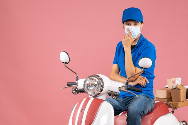 Vue de face d'un livreur confiant en masque médical portant un chapeau assis sur un scooter sur fond de pêche pastel