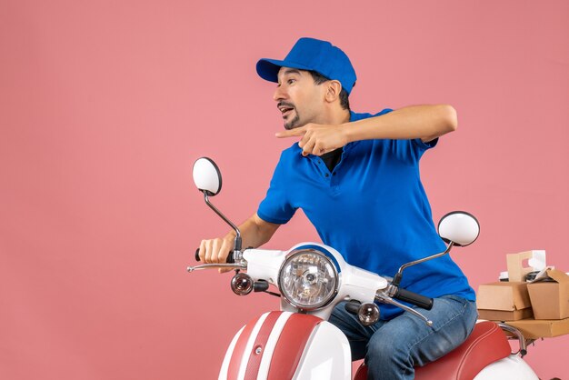 Vue de face d'un livreur curieux portant un chapeau assis sur un scooter et pointant quelque chose sur le côté droit sur fond de pêche pastel