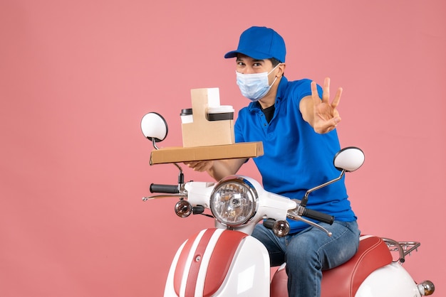 Vue de face d'un livreur masculin portant un masque portant un chapeau assis sur un scooter livrant des commandes montrant trois sur fond de pêche pastel