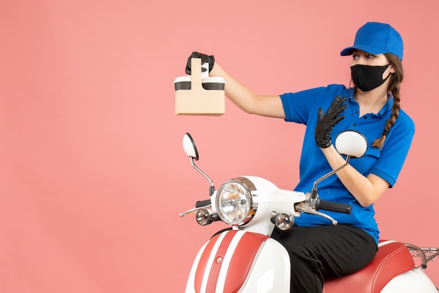 Vue de face d'un livreur plein d'espoir portant un masque médical et des gants assis sur un scooter livrant des commandes sur fond de pêche pastel