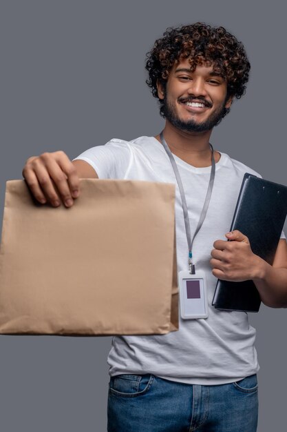 Vue de face d'un livreur satisfait avec un badge d'identification tenant un dossier et un sac en papier