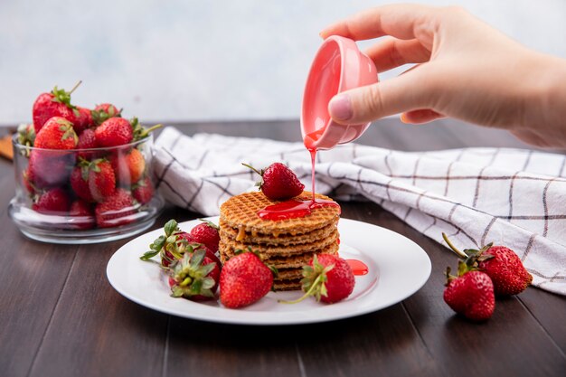 Vue de face de la main verser le sirop de fraise du bol sur les biscuits gaufres et bol de fraise sur la surface en bois