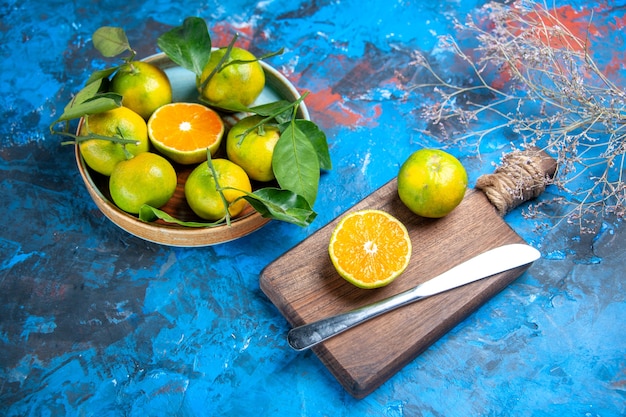 Vue de face des mandarines fraîches avec des feuilles dans un bol, un couteau de dîner à la mandarine coupé sur une planche à découper sur fond bleu