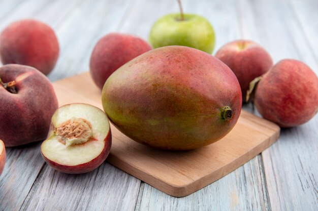 Vue de face de la mangue fraîche sur une planche de cuisine en bois avec des fruits frais tels que la pêche aux pommes sur une surface en bois gris