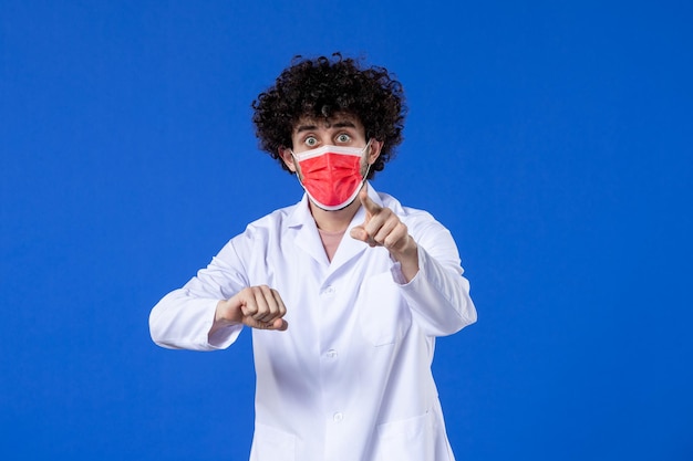 Vue de face médecin de sexe masculin en costume médical avec masque rouge sur fond bleu virus de l'hôpital covid- médecine de la santé vaccin pandémique médicaments