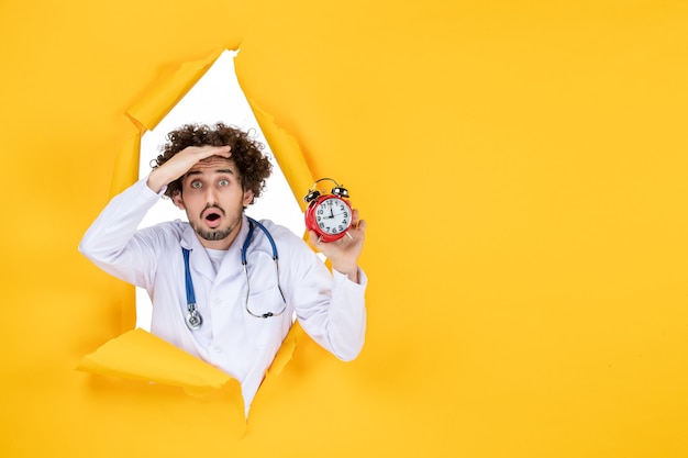 Vue de face médecin de sexe masculin en costume médical tenant des horloges sur la couleur jaune de l'hôpital medic shopping temps de médecine