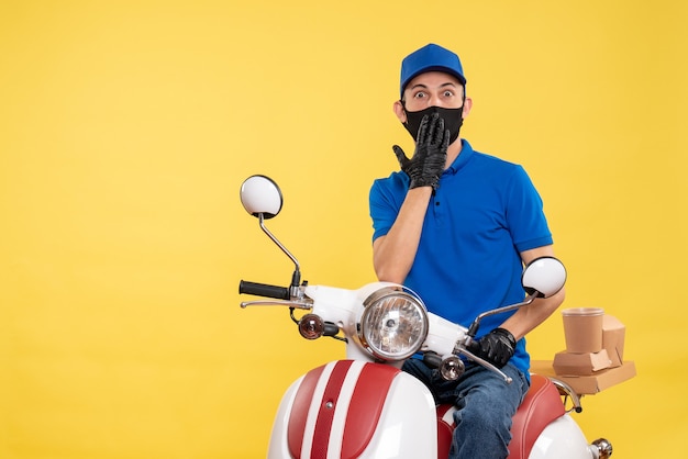 Vue de face messager masculin assis sur le vélo en masque sur le travail de livraison pandémique jaune covid- travail uniforme