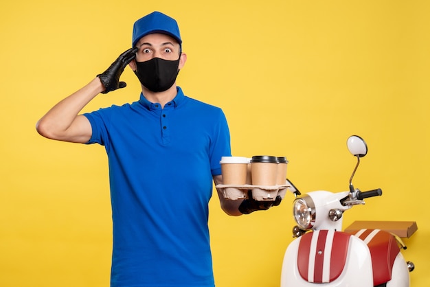 Vue de face messager masculin en masque noir tenant le café sur le travail de livraison jaune uniforme de service de travail pandémique covid