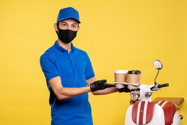 Vue de face de messager masculin en masque tenant du café sur un emploi jaune covid- uniforme de prestation de services en cas de pandémie