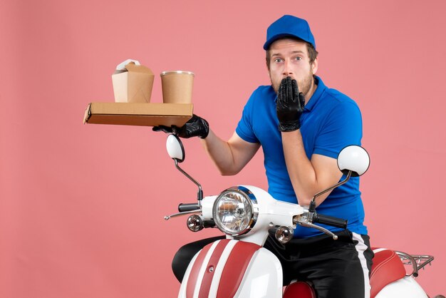 Vue de face messager masculin en uniforme bleu tenant une boîte de café et de nourriture sur la couleur du vélo de travail de livraison de restauration rapide de service rose