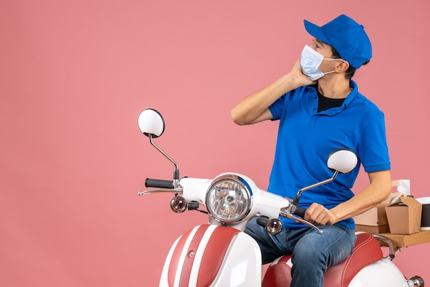 Vue de face d'un messager réfléchi en masque médical portant un chapeau assis sur un scooter sur fond de pêche pastel