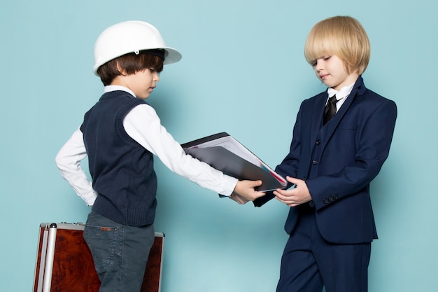 Une vue de face mignon garçon d'affaires en costume classique bleu posant tenant une valise marron-argent avec un autre garçon donnant des dossiers de mode de travail d'entreprise