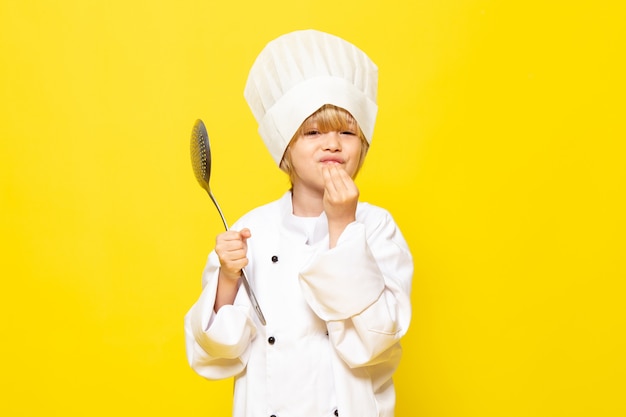 Une vue de face mignon petit enfant en costume de cuisinier blanc et chapeau de cuisinier blanc tenant une cuillère en argent sur le mur jaune enfant cuisiner des aliments de cuisine