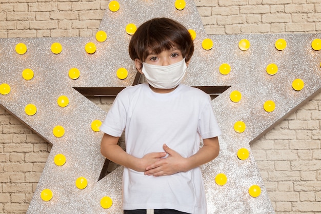 Une vue de face mignon petit enfant en t-shirt blanc jean foncé masque stérile blanc sur l'étoile conçu stand jaune et fond clair