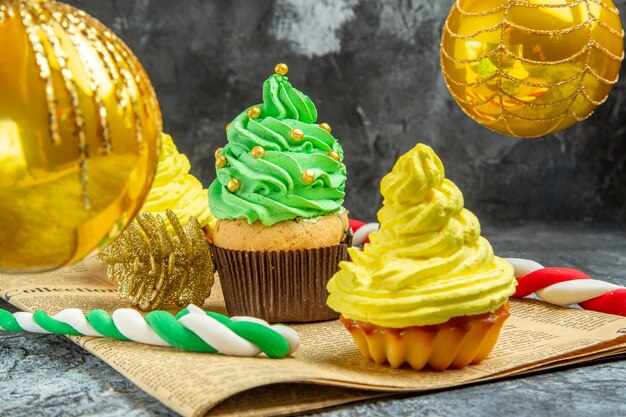 Vue de face mini cupcakes colorés boules d'arbre de Noël bonbons de Noël sur journal sur photo de Noël sombre