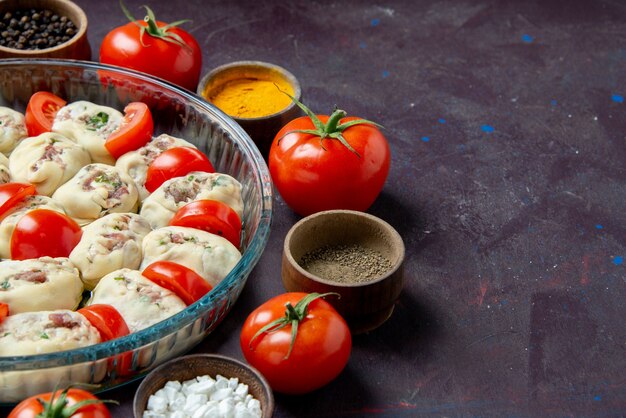 Vue de face des morceaux de pâte crue avec des assaisonnements et des tomates sur un mur sombre