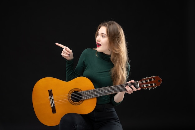 Vue de face d'une musicienne heureuse tenant une guitare et pointant quelque chose sur le côté droit sur fond noir