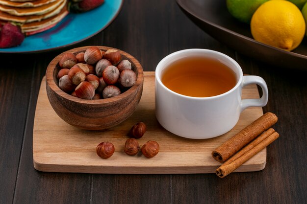 Vue de face de la noisette dans un bol avec une tasse de thé et de cannelle sur une planche sur une surface en bois