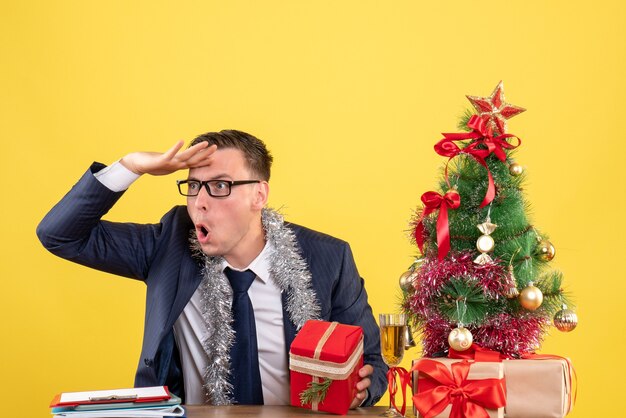 Vue de face de l'observation du jeune homme avec des lunettes assis à la table près de l'arbre de Noël et présente sur jaune