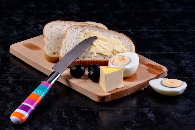 Vue de face œuf à la coque à bord avec des olives et des tranches de pain avec du beurre et un couteau