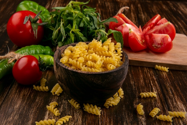 Vue de face des pâtes crues dans un bol avec des tranches de tomate en tranches sur une planche à découper avec un bouquet de menthe et de piment sur une surface en bois