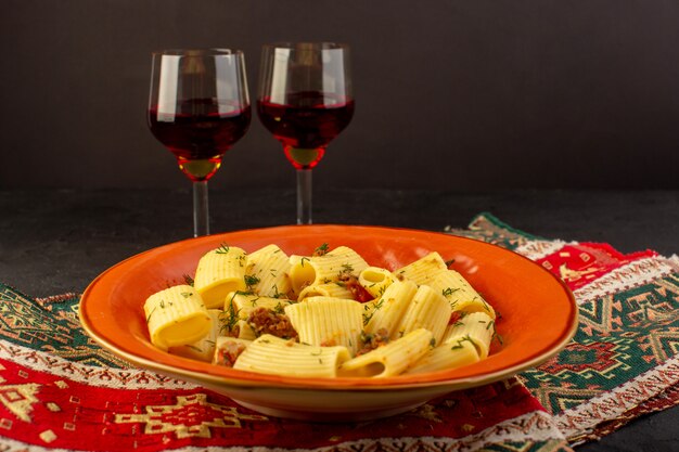 Une vue de face pâtes italiennes cuites savoureuses salées à l'intérieur de la plaque orange ronde avec des verres de vin sur un tapis conçu et un bureau sombre