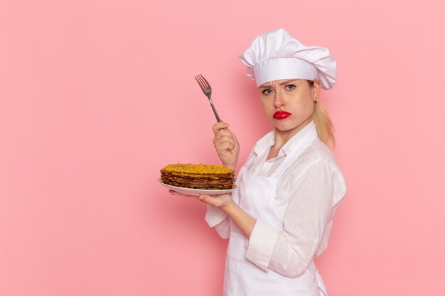 Vue de face de la pâtissière en tenue blanche tenant de délicieuses pâtisseries et une fourchette sur le mur rose pâtisserie gâteau de cuisson de confiserie