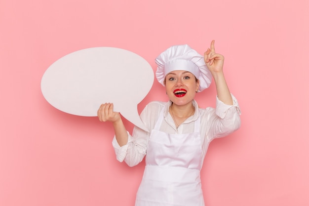 Vue de face de la pâtissière en vêtements blancs posant avec panneau blanc souriant sur le mur rose confiserie pâtisserie travail travail