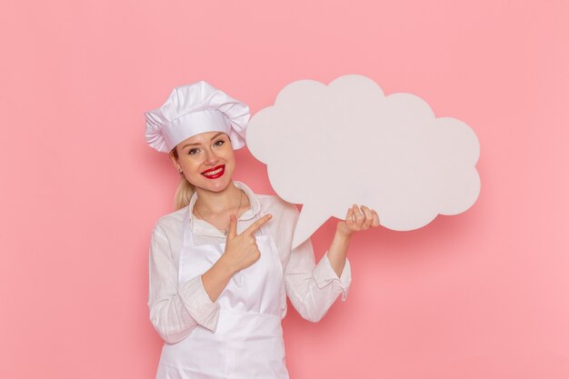 Vue de face de la pâtissière en vêtements blancs tenant grand panneau blanc sur le mur rose cuisinier travail cuisine cuisine alimentaire