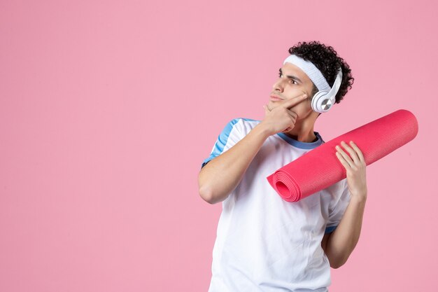 Vue de face pensant jeune homme en vêtements de sport avec tapis de yoga et écouteurs