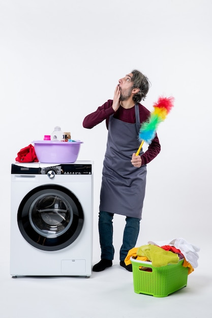 Vue de face perplexe femme de ménage tenant un plumeau debout près du panier à linge de la machine à laver sur un mur blanc