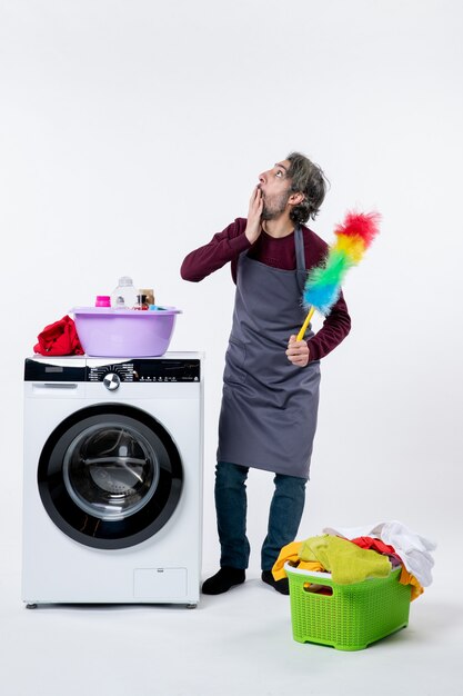 Vue de face perplexe femme de ménage tenant un plumeau debout près du panier à linge de la machine à laver sur un mur blanc